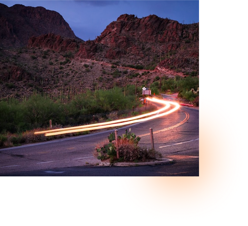 Curving desert road with light trails and rocky hills at dusk.