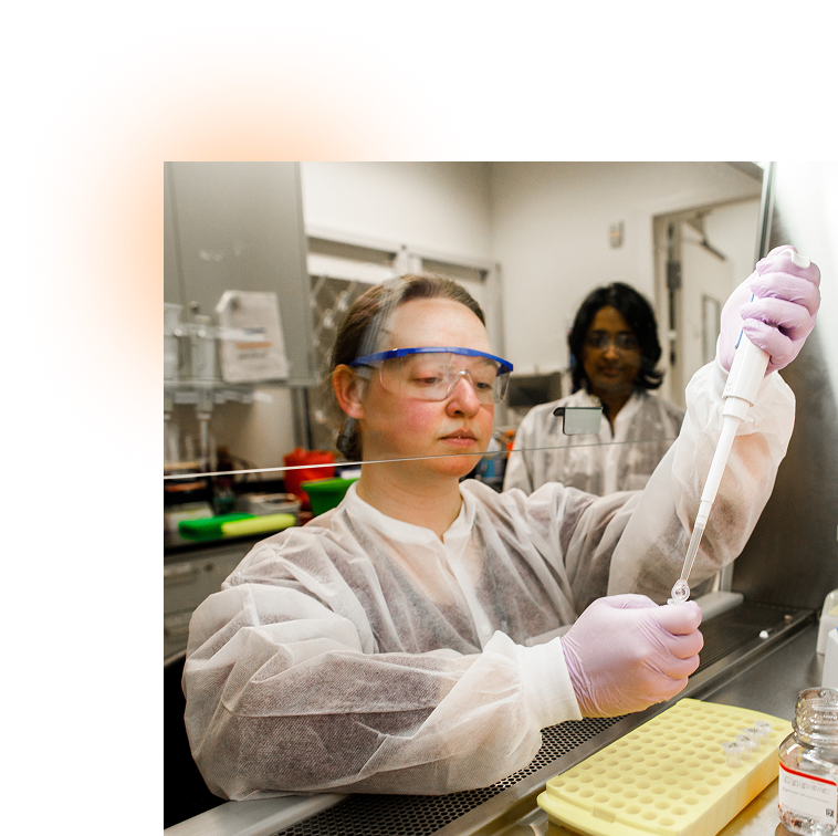A woman wearing PPE uses a pipette in a laboratory while a coworker looks on