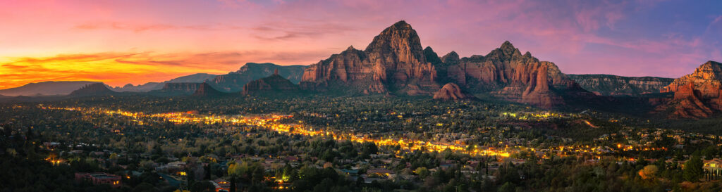 Panoramic view of Sedona, Arizona, at sunset with illuminated town, red rock formations, and vibrant sky.